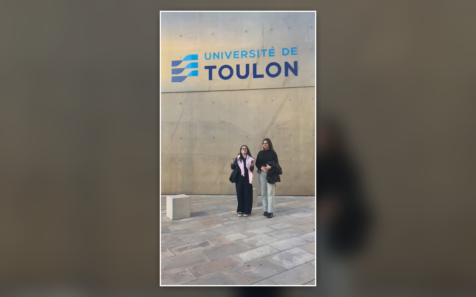 Deux étudiantes devant le mur portant le logo de l’Université de Toulon.